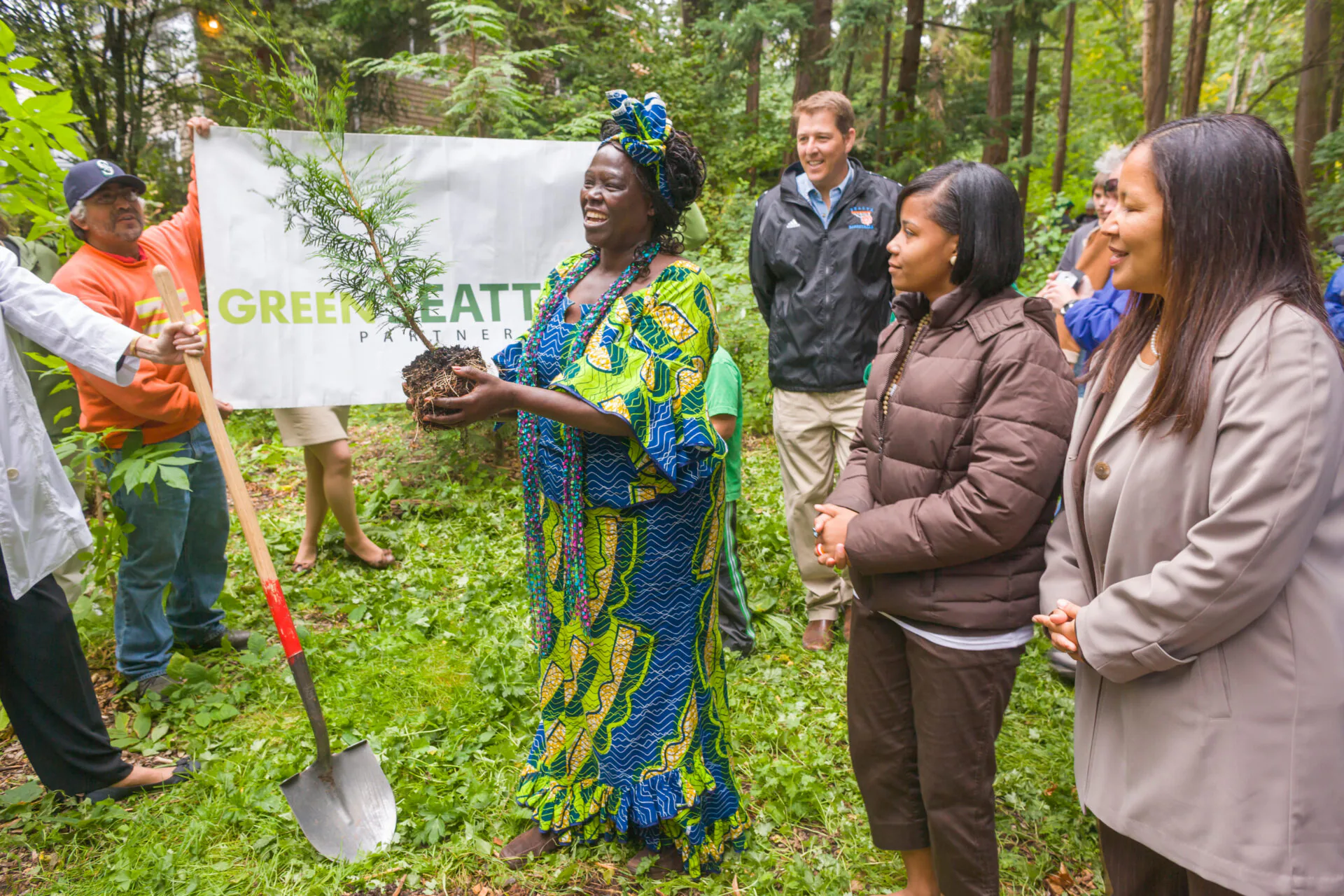 Wangari Maathai steht im Pelly Place Natural Area im Freien, umgeben von üppigen grünen Bäumen und dichter Vegetation. Sie lächelt und trägt ein bunt gemustertes Kleid mit passendem Kopftuch. In einer Hand hält sie einen jungen Baum, der kurz vor der Einpflanzung steht.